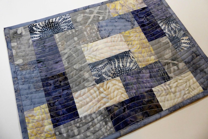 May include: A quilted table runner with a patchwork design featuring various shades of blue, grey, and white. The fabric has a subtle floral pattern.