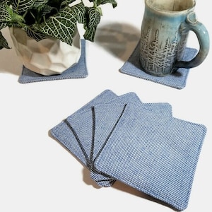May include: A collection of blue and white woven fabric coasters. One coaster supports a ceramic mug, another supports a potted plant. The coasters are square with rounded corners. The fabric has a textured appearance.