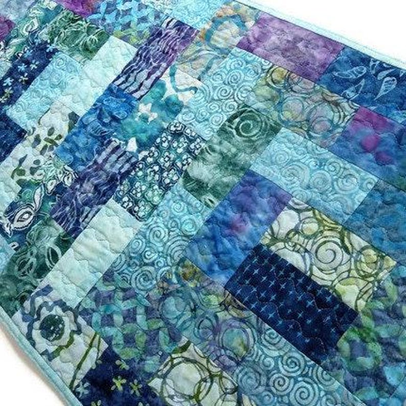 May include: A blue and green patchwork table runner with a chevron pattern. The runner is made of various fabrics with different patterns, including swirls, flowers, and geometric designs.
