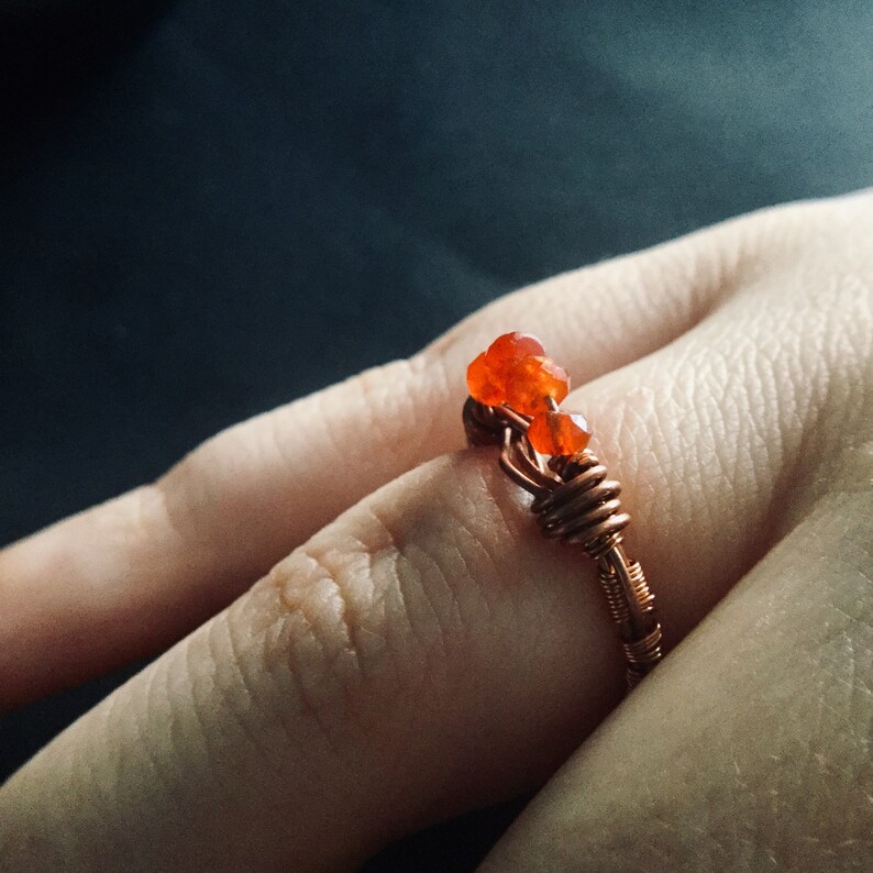 May include: A copper wire ring with three orange carnelian stones. The ring is on a person's finger.