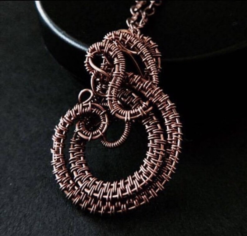K&ouml;nnte beinhalten: Eine Halskette mit einem Anh&auml;nger aus geflochtenem Kupferdraht. Der Anh&auml;nger besteht aus mehreren Schlaufen und Spiralen aus Draht, die ein einzigartiges und kompliziertes Muster bilden.