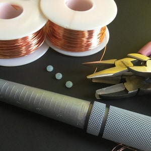 Peut inclure: Gros plan sur des fournitures de fabrication de bijoux, notamment deux bobines de fil de cuivre, quatre perles bleu clair, un ajusteur de bague et deux pinces.