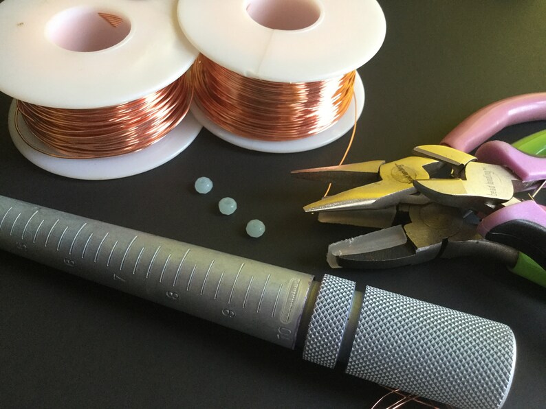 Peut inclure: Gros plan sur des fournitures de fabrication de bijoux, notamment deux bobines de fil de cuivre, quatre perles bleu clair, un ajusteur de bague et deux pinces.