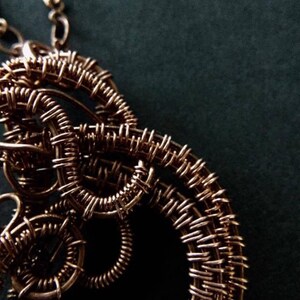 K&ouml;nnte beinhalten: Eine Halskette aus Kupferdraht mit einem gro&szlig;en, komplizierten Anh&auml;nger. Der Anh&auml;nger besteht aus mehreren Schleifen und Spiralen aus Draht, die ein komplexes und organisches Design bilden.