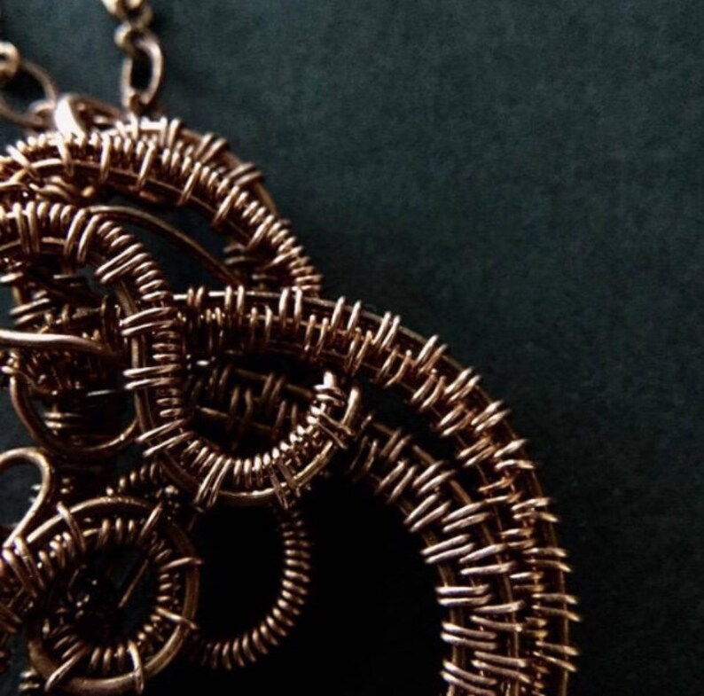 K&ouml;nnte beinhalten: Eine Halskette aus Kupferdraht mit einem gro&szlig;en, komplizierten Anh&auml;nger. Der Anh&auml;nger besteht aus mehreren Schleifen und Spiralen aus Draht, die ein komplexes und organisches Design bilden.