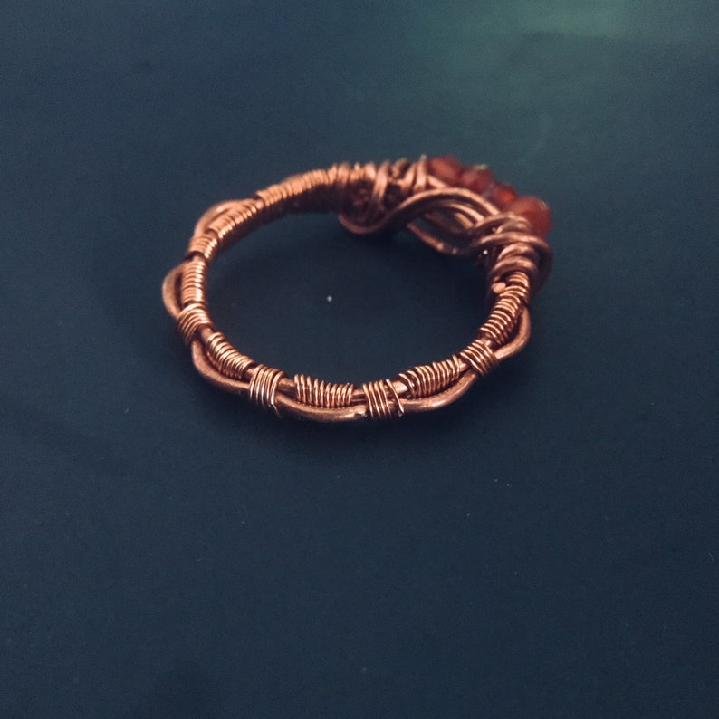 May include: A copper wire ring with three small orange stones set in the wire. The ring is on a dark blue background.