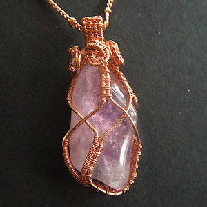 May include: A copper wire wrapped pendant featuring a large, faceted amethyst gemstone. The pendant is a delicate and intricate design with a woven wire pattern.