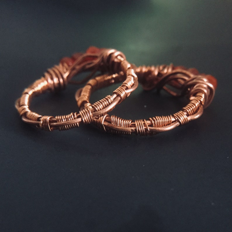 May include: Two copper wire wrapped rings with red gemstones. The rings are intertwined and the gemstones are visible.