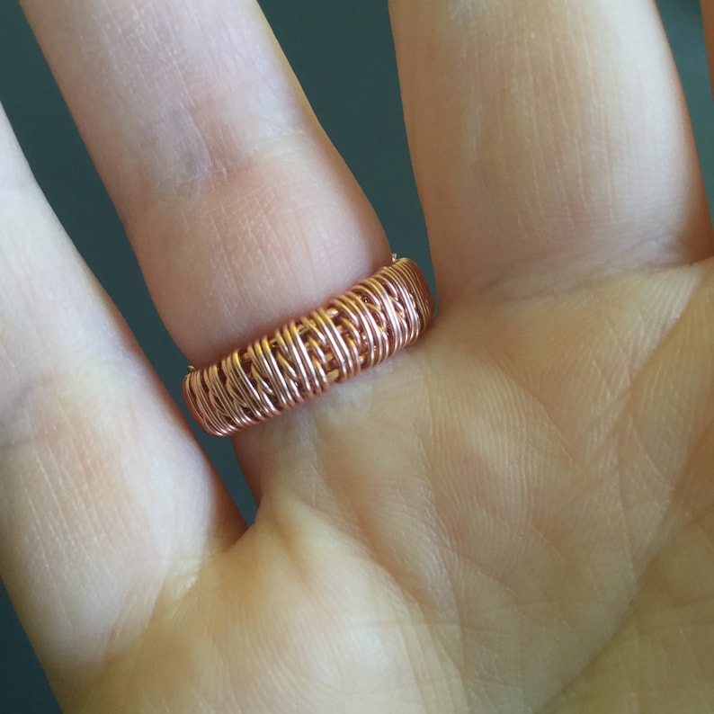 Peut inclure: Gros plan d'une bague en fil de cuivre sur le doigt d'une personne. La bague est compos&eacute;e de plusieurs brins de fil entrelac&eacute;s pour cr&eacute;er un design textur&eacute; et tress&eacute;.