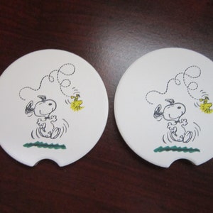 Puede incluir: Dos posavasos blancos para coche con un diseño de Snoopy saltando con un pájaro amarillo. Los posavasos tienen un recorte curvo y un diseño de hierba verde. Cada posavasos mide aproximadamente 8 cm de diámetro.