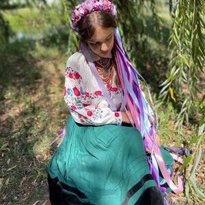 May include: A person wearing a floral crown and traditional Ukrainian clothing. The white blouse has red and blue floral embroidery. The long green skirt has a black trim. Long colorful ribbons cascade down from the floral crown.