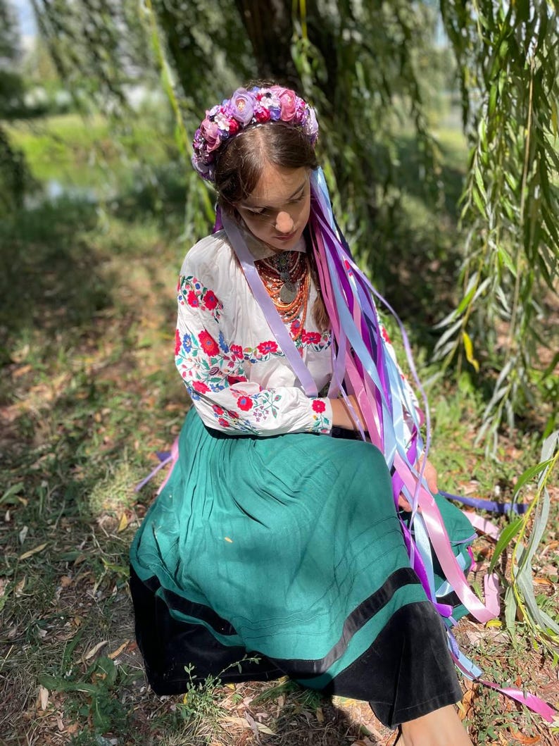 May include: A person wearing a floral crown and traditional Ukrainian clothing. The white blouse has red and blue floral embroidery. The long green skirt has a black trim. Long colorful ribbons cascade down from the floral crown.