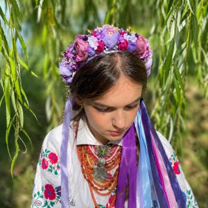 May include: A floral crown with purple and pink flowers adorns a person's head. Long, colorful ribbons cascade down, complementing the white embroidered shirt with red and blue floral patterns. A necklace with orange beads and a silver pendant adds detail.