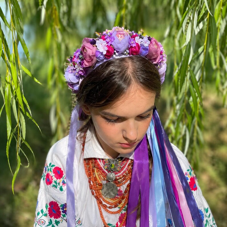 May include: A floral crown with purple, pink, and lavender flowers. The crown sits atop a person's head, with long, colorful ribbons cascading down. The person is wearing a white embroidered shirt and a beaded necklace. The background is a blurred green.