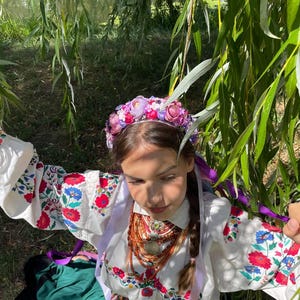 May include: A young person wearing a traditional white embroidered blouse with red and blue floral patterns, a green skirt, and a floral crown. The person is also wearing a layered necklace with a pendant and purple ribbons.