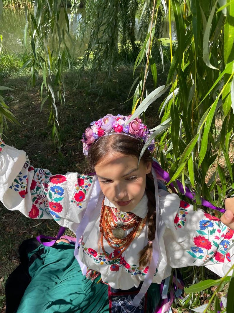 May include: A young person wearing a traditional white embroidered blouse with red and blue floral patterns, a green skirt, and a floral crown. The person is also wearing a layered necklace with a pendant and purple ribbons.