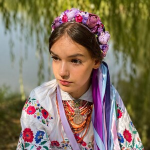 May include: A young person wearing a floral crown with purple, pink, and white flowers. They are also wearing a white embroidered shirt with red and blue floral patterns, and a necklace with a large pendant and orange beads. Long purple and blue ribbons hang down.