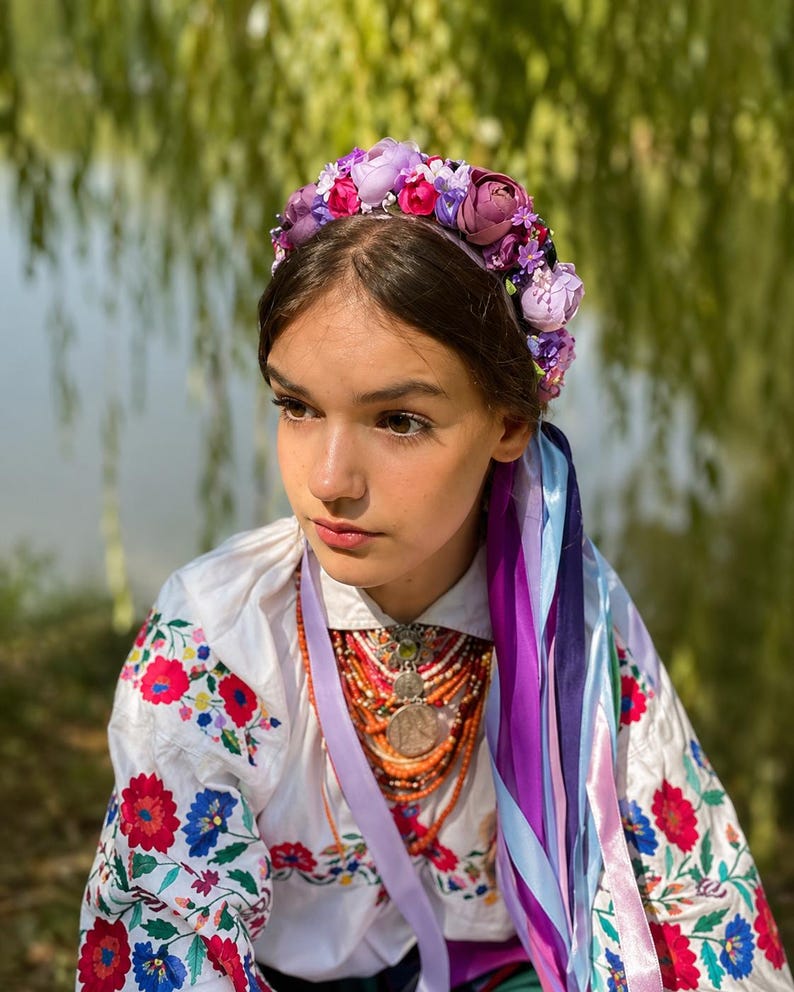 May include: A young person wearing a floral crown with purple, pink, and white flowers. They are also wearing a white embroidered shirt with red and blue floral patterns, and a necklace with a large pendant and orange beads. Long purple and blue ribbons hang down.