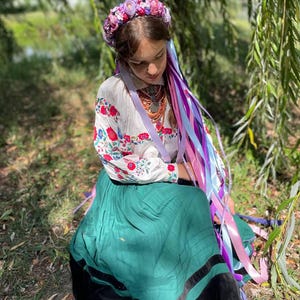 May include: A person wearing a floral crown and traditional embroidered blouse with a green skirt. The blouse has red and blue floral embroidery. Long, colorful ribbons cascade down from the crown.