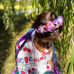 May include: A floral crown with a variety of pink, purple, and blue flowers. The crown is worn with a white embroidered blouse with red and blue floral patterns and colorful ribbons.