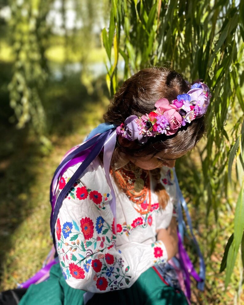 May include: A floral crown with a variety of pink, purple, and blue flowers. The crown is worn with a white embroidered blouse with red and blue floral patterns and colorful ribbons.