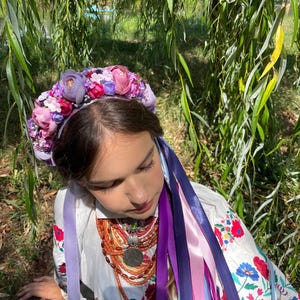 May include: A floral crown with purple and pink flowers, worn with a white embroidered blouse and colorful ribbons. The blouse features red and blue floral embroidery. A multi-strand necklace with a large pendant is also visible.