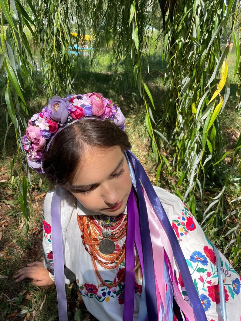 May include: A floral crown with purple and pink flowers, worn with a white embroidered blouse and colorful ribbons. The blouse features red and blue floral embroidery. A multi-strand necklace with a large pendant is also visible.