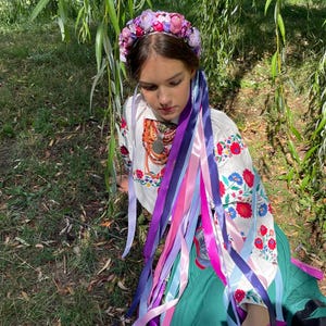 May include: A woman wearing a floral crown and a white embroidered blouse with colorful floral patterns. Long, colorful ribbons cascade down from the crown and blouse, adding a vibrant touch. The woman is sitting in a grassy area.