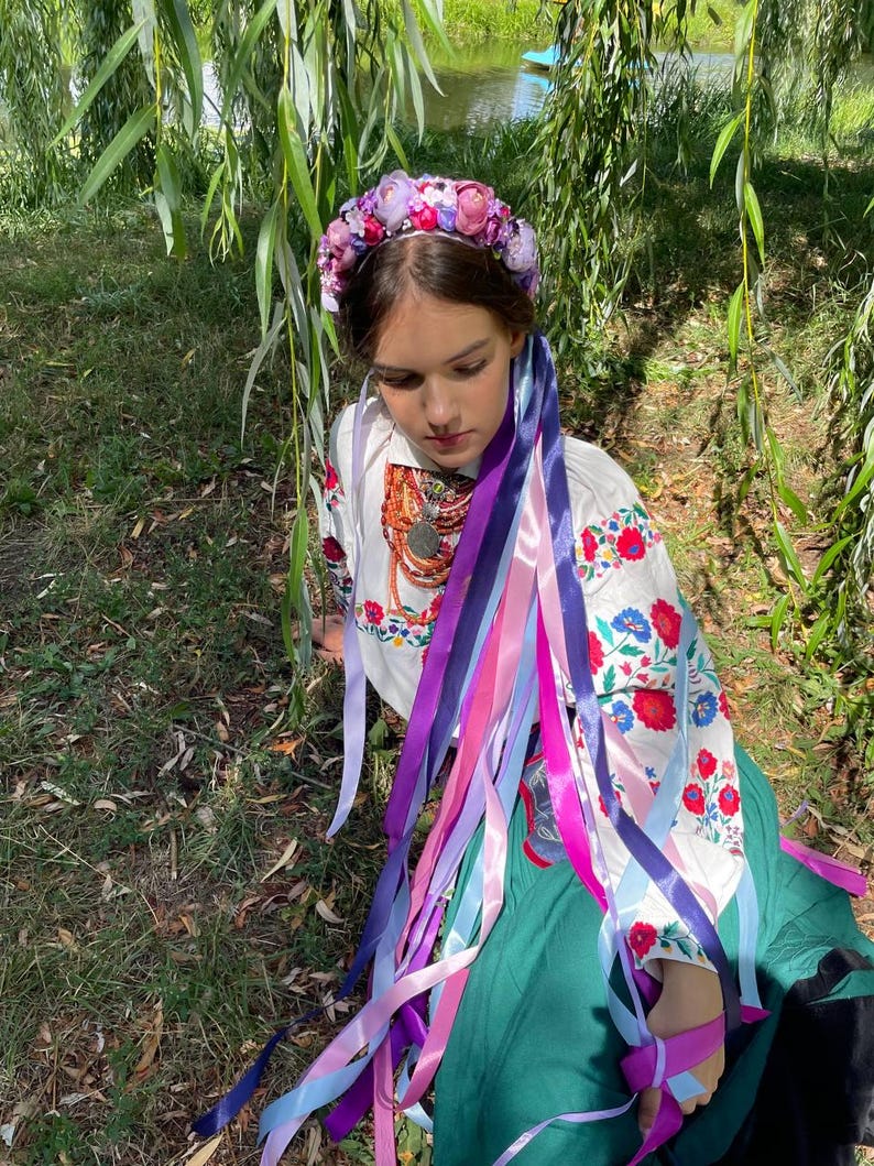 May include: A woman wearing a floral crown and a white embroidered blouse with colorful floral patterns. Long, colorful ribbons cascade down from the crown and blouse, adding a vibrant touch. The woman is sitting in a grassy area.