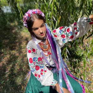 May include: A woman wearing a traditional Ukrainian outfit. The white blouse features red and blue floral embroidery on the sleeves. She wears a floral crown and long colorful ribbons. A green skirt and beaded necklace complete the ensemble.