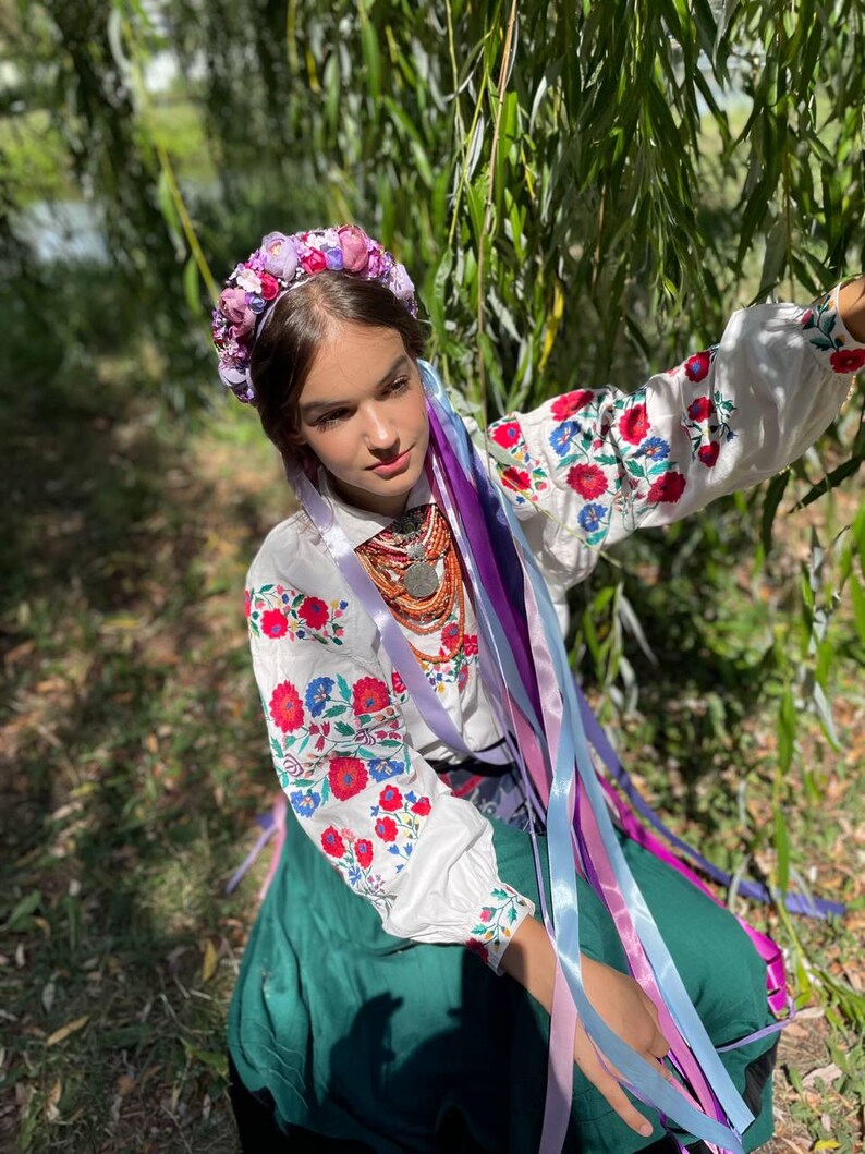 May include: A woman wearing a traditional Ukrainian outfit. The white blouse features red and blue floral embroidery on the sleeves. She wears a floral crown and long colorful ribbons. A green skirt and beaded necklace complete the ensemble.