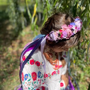 May include: A floral crown with pink, purple, and blue flowers. The crown sits atop a person's head, with long, colorful ribbons cascading down. The person is wearing a white embroidered blouse with red and blue floral patterns.