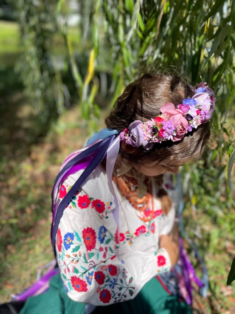 May include: A floral crown with pink, purple, and blue flowers. The crown sits atop a person's head, with long, colorful ribbons cascading down. The person is wearing a white embroidered blouse with red and blue floral patterns.