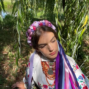 May include: A floral crown with purple and pink flowers adorns the head of a person wearing a white embroidered shirt. Long, colorful ribbons cascade down. A necklace with a large pendant and orange beads is also visible.