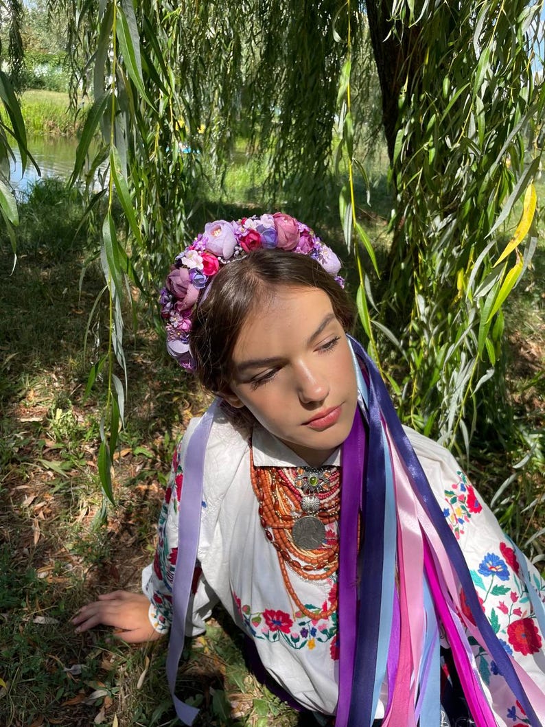 May include: A floral crown with purple and pink flowers adorns the head of a person wearing a white embroidered shirt. Long, colorful ribbons cascade down. A necklace with a large pendant and orange beads is also visible.