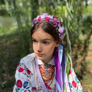 May include: A young person wearing a floral crown with purple, pink, and red flowers. They are wearing a white embroidered shirt with red and blue floral patterns. Colorful ribbons hang from the crown.