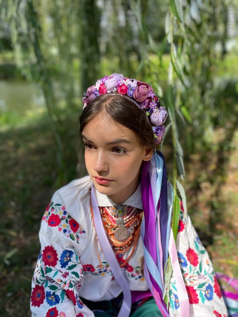May include: A young person wearing a floral crown with purple, pink, and red flowers. They are wearing a white embroidered shirt with red and blue floral patterns. Colorful ribbons hang from the crown.