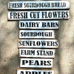 Puede incluir: Una colecci&oacute;n de letreros de madera r&uacute;sticos con fondos pintados de blanco y letras negras. Los letreros muestran frases como "Fresh Sourdough Bread", "Fresh Cut Flowers" y "Farm Stand", sugiriendo un mercado de agricultores.