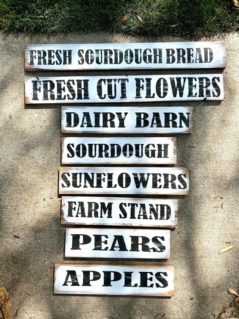 Puede incluir: Una colecci&oacute;n de letreros de madera r&uacute;sticos con fondos pintados de blanco y letras negras. Los letreros muestran frases como "Fresh Sourdough Bread", "Fresh Cut Flowers" y "Farm Stand", sugiriendo un mercado de agricultores.