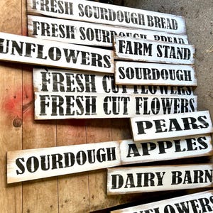 Puede incluir: Un conjunto de letreros de madera r&uacute;sticos, pintados de blanco con letras negras. Los letreros muestran frases como "Fresh Sourdough Bread", "Farm Stand", "Sunflowers" y "Pears". Los letreros est&aacute;n dispuestos sobre una superficie de madera.