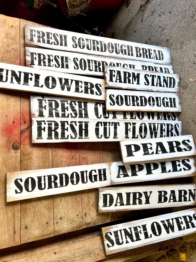 Puede incluir: Un conjunto de letreros de madera r&uacute;sticos, pintados de blanco con letras negras. Los letreros muestran frases como "Fresh Sourdough Bread", "Farm Stand", "Sunflowers" y "Pears". Los letreros est&aacute;n dispuestos sobre una superficie de madera.