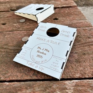 May include: A white wooden game with a circular hole and the text "Girls Drink" and "Guys Drink". The game also has the text "MAKE A RULE" and "Mr. & Mrs Newton 2025".
