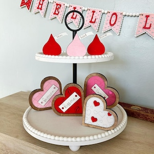 May include: A two-tiered white tiered tray with a black stand. The top tier has three red, pink, and white heart-shaped decorations with the words "Mom", "Isabel", and "Emma" on them. The bottom tier has four wooden heart-shaped decorations with the words "Love", "Kiss Me", "Bemine", and a heart pattern.