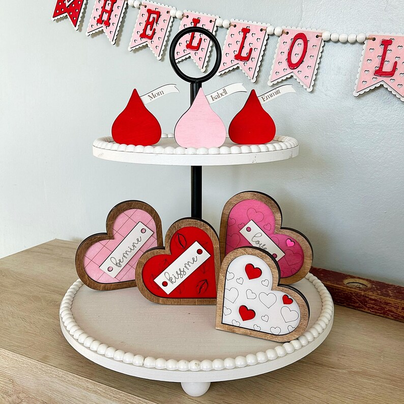 May include: A two-tiered white tiered tray with a black stand. The top tier has three red, pink, and white heart-shaped decorations with the words "Mom", "Isabel", and "Emma" on them. The bottom tier has four wooden heart-shaped decorations with the words "Love", "Kiss Me", "Bemine", and a heart pattern.