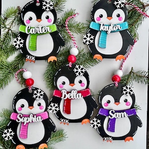 May include: Five black and white penguin ornaments featuring orange feet, pink cheeks, and colorful scarves. Each penguin is personalized with names including Carter, Taylor, Bella, Sophia, and Sam, along with the year 2026 and white snowflake accents.