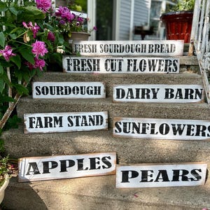 Puede incluir: Una colecci&oacute;n de letreros r&uacute;sticos de madera con letras negras sobre fondo blanco. Los letreros muestran varios productos frescos de la granja, incluyendo "Fresh Sourdough Bread", "Fresh Cut Flowers", "Apples" y "Pears".