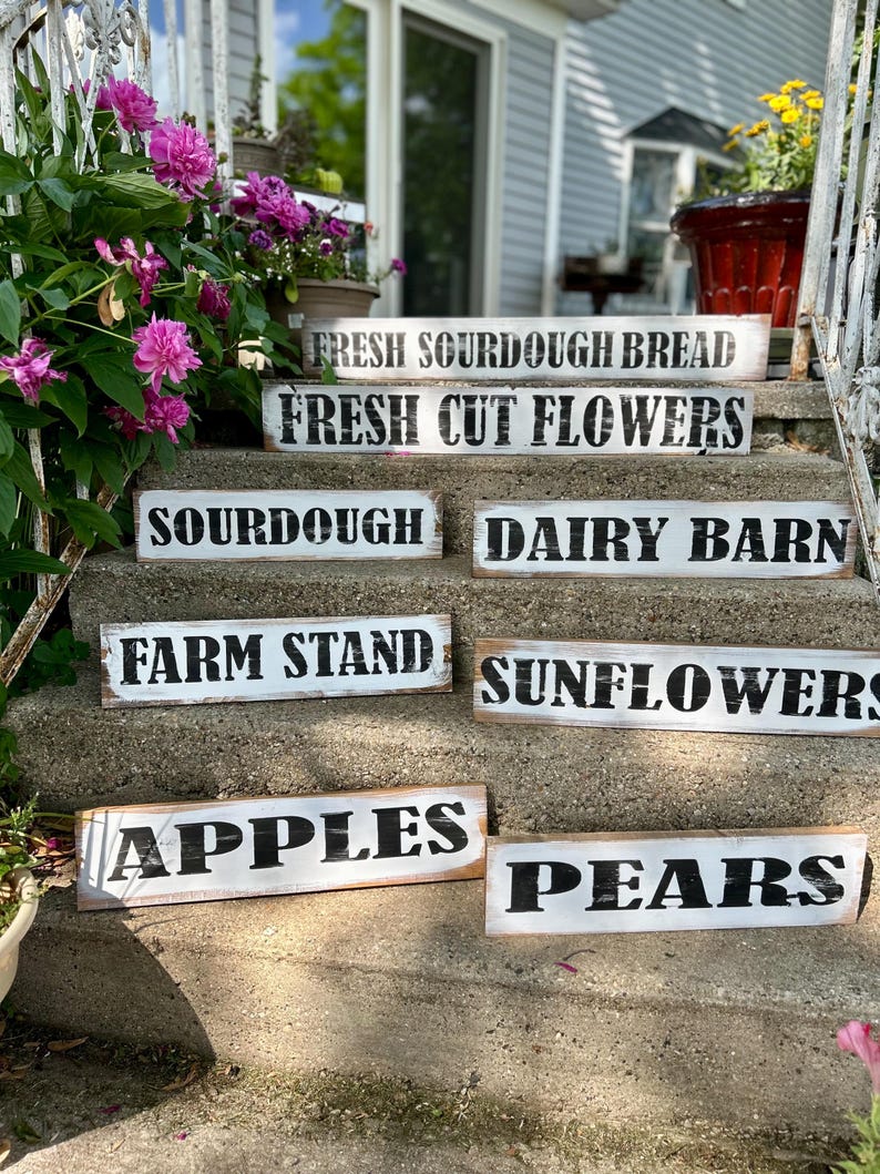 Puede incluir: Una colecci&oacute;n de letreros r&uacute;sticos de madera con letras negras sobre fondo blanco. Los letreros muestran varios productos frescos de la granja, incluyendo "Fresh Sourdough Bread", "Fresh Cut Flowers", "Apples" y "Pears".