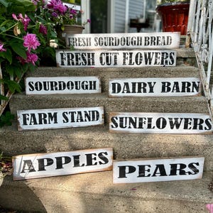 Puede incluir: Una colecci&oacute;n de letreros de madera rectangulares r&uacute;sticos con fondos blancos y letras negras. Los letreros muestran frases como "Fresh Sourdough Bread", "Fresh Cut Flowers", "Apples" y "Pears", lo que sugiere un tema de mercado agr&iacute;cola.