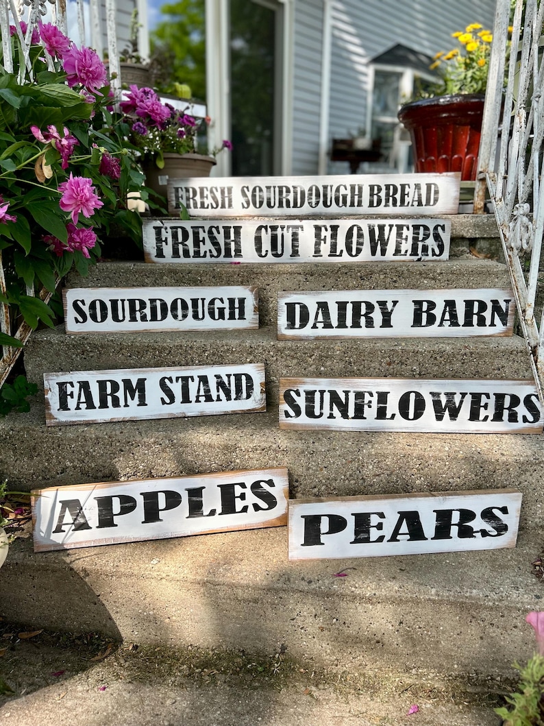 Puede incluir: Una colecci&oacute;n de letreros de madera rectangulares r&uacute;sticos con fondos blancos y letras negras. Los letreros muestran frases como "Fresh Sourdough Bread", "Fresh Cut Flowers", "Apples" y "Pears", lo que sugiere un tema de mercado agr&iacute;cola.