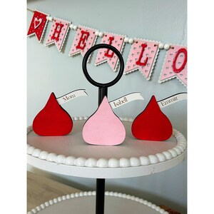 May include: Three wooden chocolate kiss-shaped ornaments with personalized names on white ribbon banners. The ornaments are red, pink, and red and are displayed on a white tiered cake stand with a black base. The background features a red and white "Hello" banner.